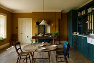 A lovely modern kitchen that retains the charm of its original structure and flooring, using the three primary colours in a muted, mature palette. The spacious kitchen features a Clerkenwell Blue full-height pantry cabinet, a long sink run with a marble worktop, and an open countertop cabinet and shelf. An emerald green stove sits within the chimney breast, accompanied by a deVOL cream pendant light above a vintage round wooden table, with matching chairs and a prep table.