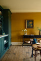 A lovely modern kitchen that retains the charm of its original structure and flooring, using the primary colours in a muted, mature palette. The spacious room features a Clerkenwell Blue full-height pantry cabinet, a long sink run with a marble worktop, and an open countertop cabinet with a shelf. At the back of the kitchen, a vintage black metal console table with drawers sits against a warm orange-yellow wall, adorned with vases of flowers and a gold-framed, detailed still life painting.