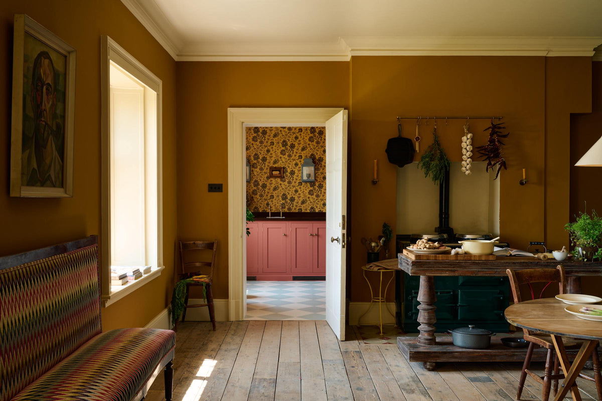 Looking through the door into a lovely modern kitchen—one that retains the charm of its original structure and flooring, and uses primary colours in a muted, mature palette—you’ll see light red sink run with a polished sapele worktop, a single sink with aged brass Mayan taps, strikingly detailed floral wallpaper, and light blue and white porcelain floor tiles.