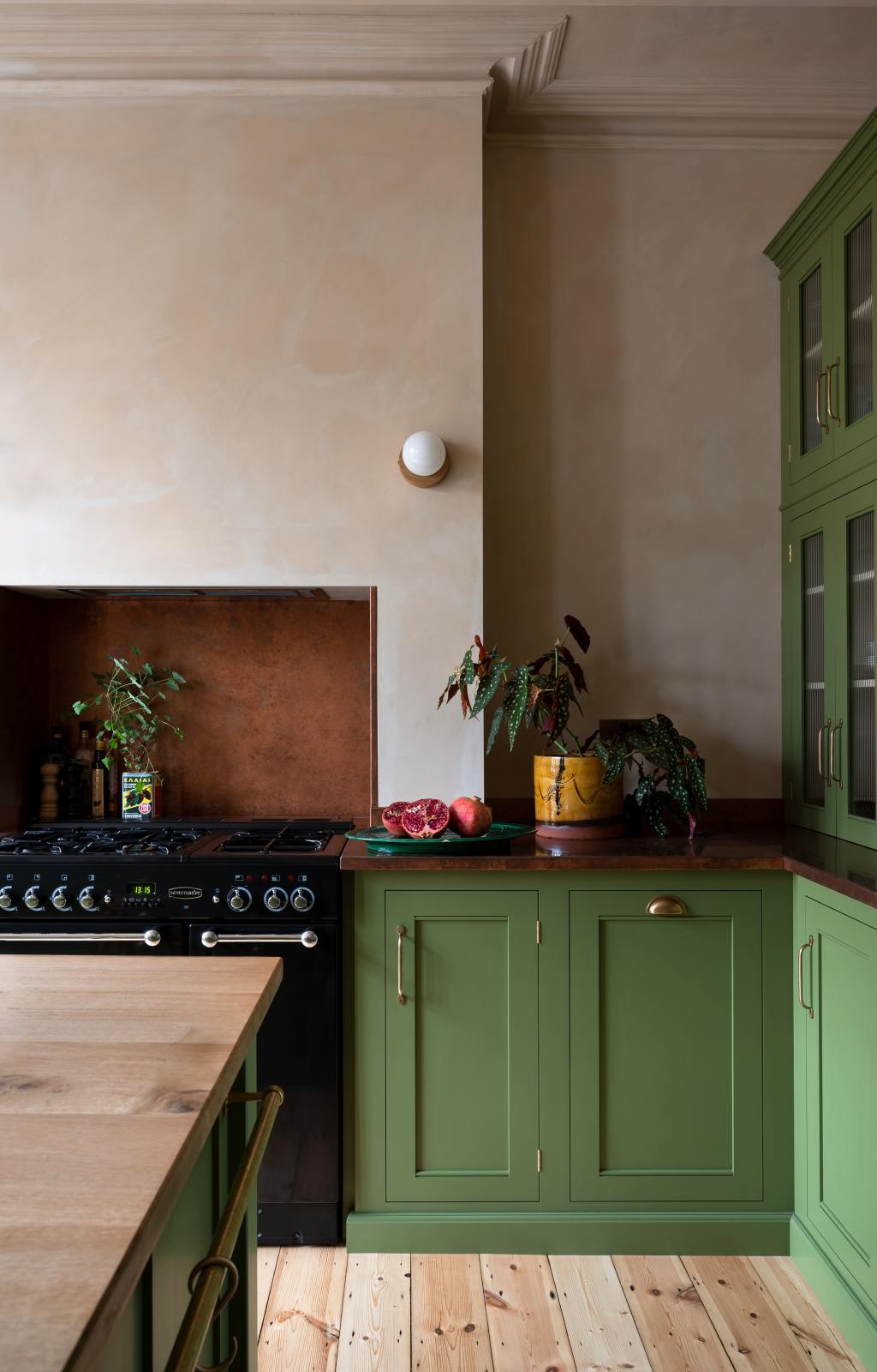 A closer look at a romantic, decorative, Parisian-inspired green classic English kitchen, featuring an L-shaped sink and cooker run with an aged copper worktop and an oak worktop kitchen island. Adorned with lush green house plants, and a strikingly red plate of grapefruits with brown, marble-washed walls and ceiling.