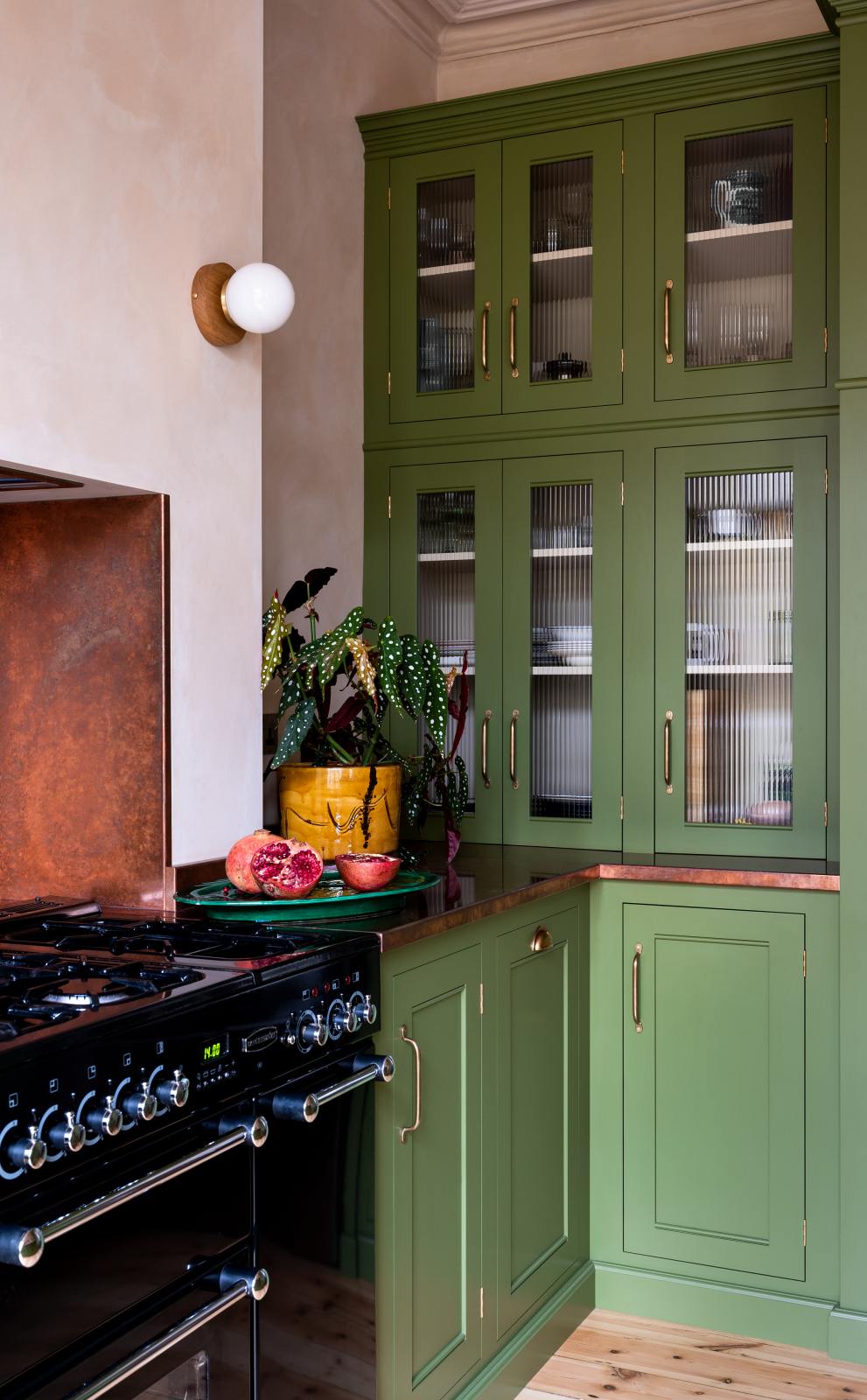 A closer look at a romantic, decorative, Parisian-inspired green classic English kitchen, featuring an L-shaped sink and cooker run with an aged copper worktop, black gas stove cosied in the chimney breast with a copper splashback, and glazed reeded counter cupboards. Adorned with lush green house plants, and a strikingly red plate of grapefruits with brown marble wash walls and ceiling.