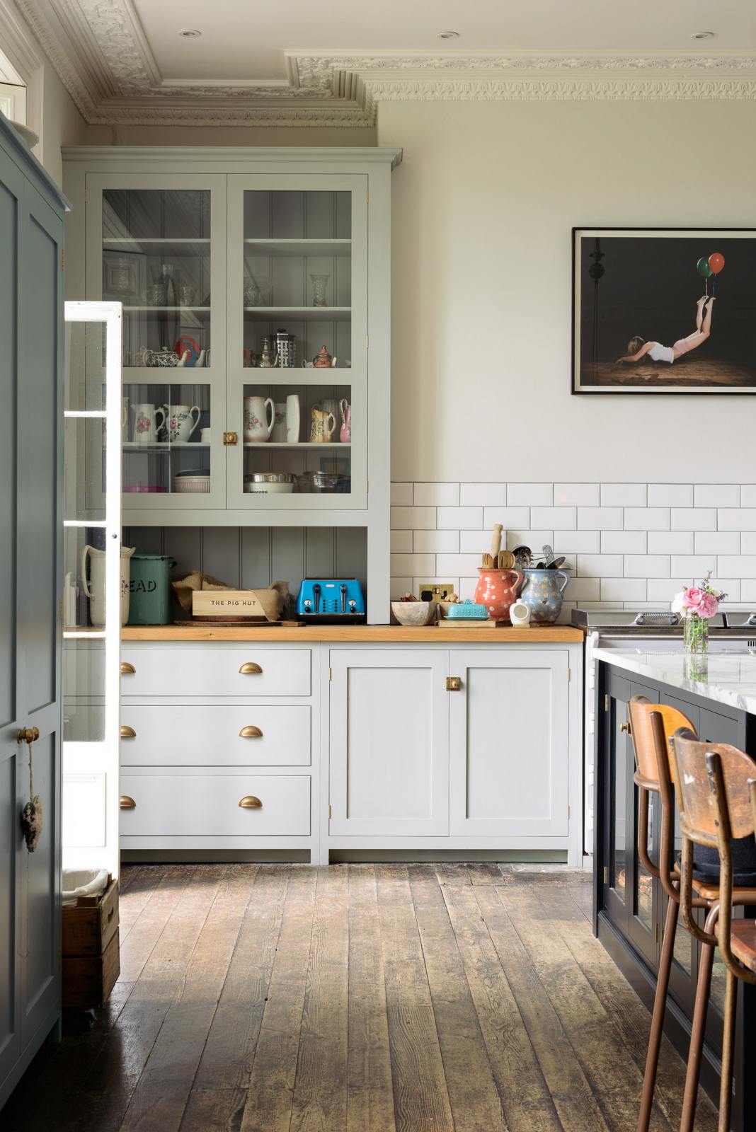 A closer look at a tall-ceilinged classic English kitchen centres around a wide painter’s black island with an aged brass Ionian tap, marble worktop, and glazed base cabinets. A cosy breakfast nook with vintage stools sits opposite an off-white cooker run and matching wall cabinets. The space feels artfully lived-in—funky paintings, and kitchen essentials tell the story of a creative, full-of-life family.