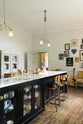 An oblique view of a tall-ceilinged classic English kitchen centres around a wide painter’s black island with an aged brass Ionian tap, marble worktop, glazed base cabinets, and a cosy breakfast nook with vintage stools. The space feels artfully lived-in—funky paintings, a guitar, and kitchen essentials tell the story of a creative, full-of-life family.