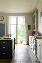 One side of a French door is left open to the lush green garden, allowing bright white natural light to pour into the tall-ceilinged, classic English kitchen centred around a wide painter’s black island with a marble worktop, and an off-white cooker run with matching wall cabinets and white porcelain splashback tiles.