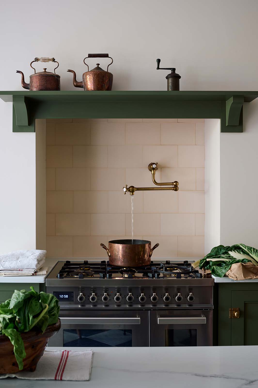 A closer look at a spacious and elegant Wilkes green classic English kitchen featuring the gas stove in the chimney breast. Adorned with an aged brass pot filler tap, cream subway splashback tiles, a copper pot, and luscious green vegetables, it embodies a classic and stylish aesthetic.