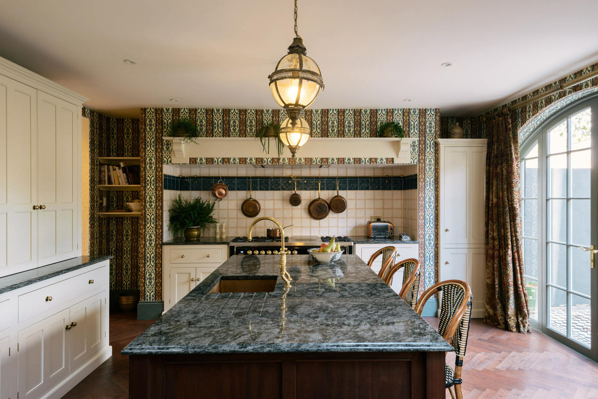 This grand and luxurious classic English kitchen features a cooker run set into the chimney breast, a large kitchen island topped with Volga blue granite, a single copper sink, an aged brass Ionian tap, and free-standing cupboards. Lavishly decorated with intricately patterned wallpaper, a William Morris pattern curtain, reclaimed splashback tiles, and hanging lantern pendant lights, it creates a royal yet comfortable and quirky space.