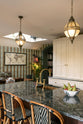 An oblique view of a grand and luxurious classic English kitchen features a large kitchen island topped with Volga blue granite, a single copper sink, an aged brass Ionian tap, and free-standing cupboards. Lavishly decorated with intricately patterned wallpaper, an artisan tea set, a vintage world map, a bowl of fruits, and hanging lantern pendant lights, it creates a royal yet comfortable and quirky space.
