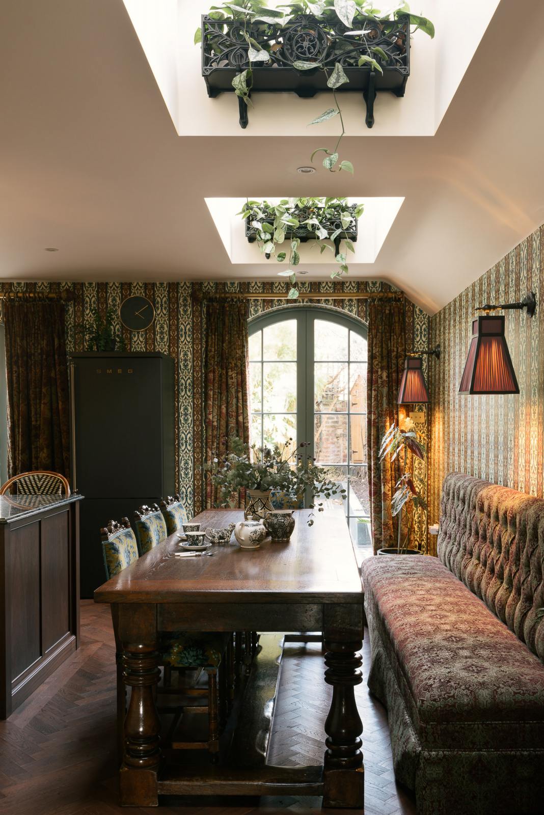 Adjacent to the grand and luxurious classic English kitchen is a dining area featuring a large solid oak table, floral-patterned chairs, and a cushioned bench. Lavishly decorated with intricately patterned wallpaper, an artisan tea set, frilled wall lamps, a large black fridge, and a unique house plant, the space creates a royal yet comfortable and quirky atmosphere.