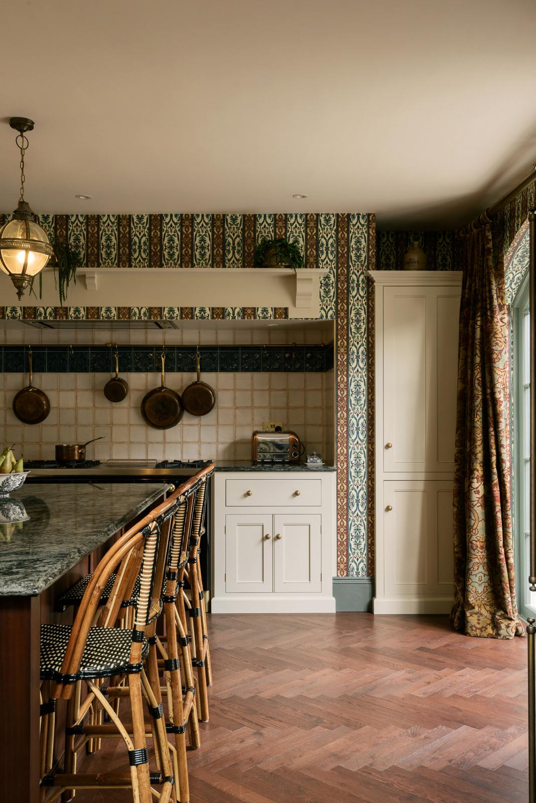 This grand and luxurious classic English kitchen features a cooker run set into the chimney breast, a large kitchen island topped with Volga blue granite, a single copper sink, an aged brass Ionian tap, and free-standing cupboards. Lavishly decorated with intricately patterned wallpaper, a William Morris pattern curtain, reclaimed splashback tiles, and hanging lantern pendant lights, it creates a royal yet comfortable and quirky space.