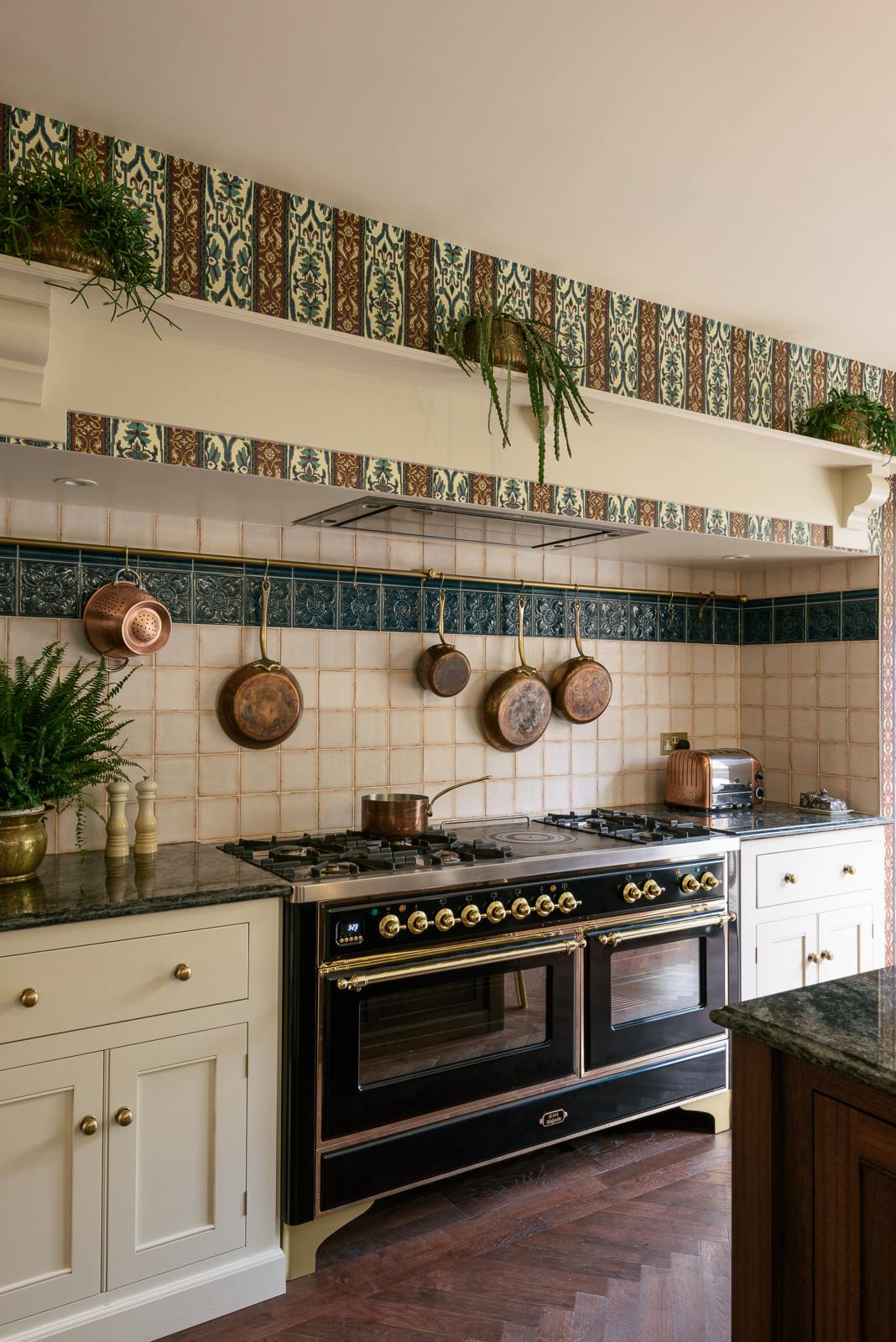 This close-up of a grand and luxurious classic English kitchen showcases the cooker run set into the chimney breast, topped with Volga blue granite. It features a shelf and a large black gas cooker. Lavishly decorated with intricately patterned wallpaper, reclaimed splashback tiles, and brass pots, pans, and a toaster, it creates a royal yet comfortable and quirky space.