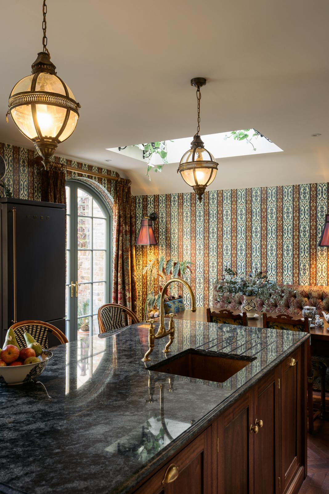 An oblique view of a grand and luxurious classic English kitchen features a large kitchen island topped with Volga blue granite, a single copper sink, an aged brass Ionian tap, and free-standing cupboards. Lavishly decorated with intricately patterned wallpaper, an artisan tea set, frilled wall lights, a bowl of fruits, and hanging lantern pendant lights, it creates a royal yet comfortable and quirky space.