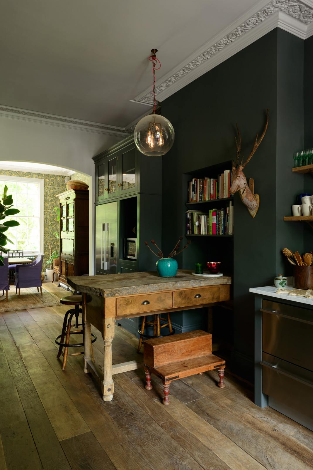 An eclectic, navy blue, classic English kitchen features a freestanding cabinet with a large silver fridge and glazed cabinets. The long, corridor-like space is beautifully decorated with beloved vintage items and furniture, paintings, a mixed-media deer wall hanging and unique lighting, giving the room a lot of whimsy, character and functionality.
