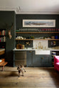 An eclectic, navy blue, classic English kitchen features a short and humble sink run with a double Belfast sink, an aged brass Ionian tap, a Carrara marble worktop, and a square splashback at the centre. The long, corridor-like space is beautifully decorated with beloved vintage items and furniture, a mixed-media deer wall hanging, unique kitchen equipment and a cute dog, giving the room a lot of whimsy, character and functionality.