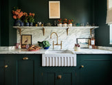 This charming Bakehouse green English kitchen features an Arabescato worktop, splashback, and counter shelf, along with a single fluted Belfast sink and an aged brass Ionian tap. Decorated with colourful bric-a-brac, framed illustrations, pretzels, washed lettuce in a blue strainer, and red chicory in a large mug, it perfectly blends functionality with personality.