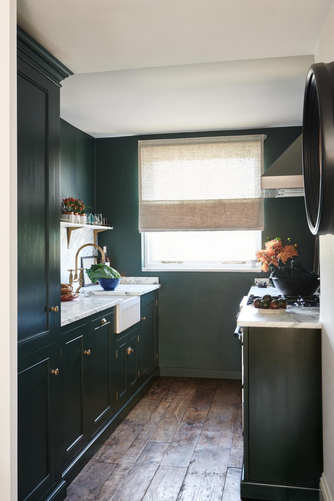A small but luxurious Bakehouse green classic English kitchen features a sink and cooker run with an Arabescato worktop, splashback and counter shelf, a single fluted Belfast sink and a full-height cabinet. It’s adorned with all sorts of colourful and intricate bric-a-brac, framed illustrations, a plate of pretzels, washed lettuce, and peachy yellow flowers, giving the kitchen functionality and personality.