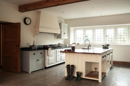 This simple and spacious classic English kitchen features a cooker run with a black granite worktop, a wall cupboard, and a kitchen island equipped with a double Belfast sink and an antique silver Ionian tap atop a solid oak worktop. Adorned with a vintage scale, a metal basket, simple ceramics, a pickling jar, red onions, and boots, it creates a humble and functional family kitchen.