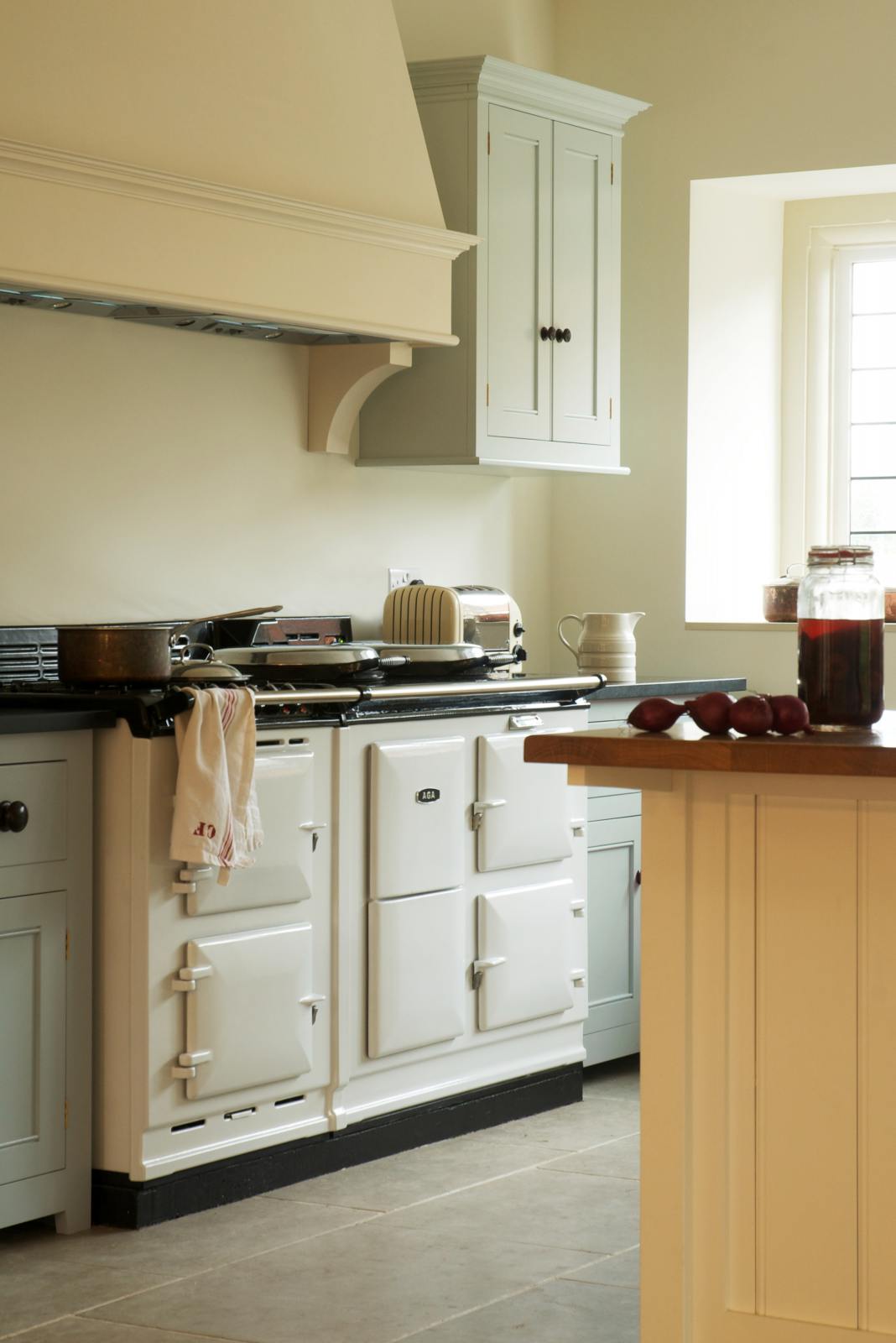An oblique view of a simple and spacious classic English kitchen features a cooker run with a black granite worktop, a wall cupboard, and a kitchen island with a solid oak worktop. Adorned with simple ceramics, a pickling jar, red onions, and a large white gas cooker, it creates a humble and functional family kitchen.