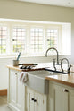 An oblique view of a simple and spacious classic English kitchen features a kitchen island with a double Belfast sink and an antique silver Ionian tap atop a solid oak worktop. Adorned with simple ceramics, a backing tray, tea towels, and a rolling pin, it creates a humble and functional family kitchen.