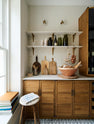 A large Haberdasher’s walk-in pantry, featuring Tambour base cabinets, open drawers, glazed countertop cupboards, a Silestone worktop, and shelves. Adorned with natural materials such as white porcelain splashback tiles, classic brass shelf brackets, an intricately patterned tiled floor, wooden chopping boards, vintage ceramics, vegetables, and loaves of bread, it makes spectacular use of the space while remaining down-to-earth.