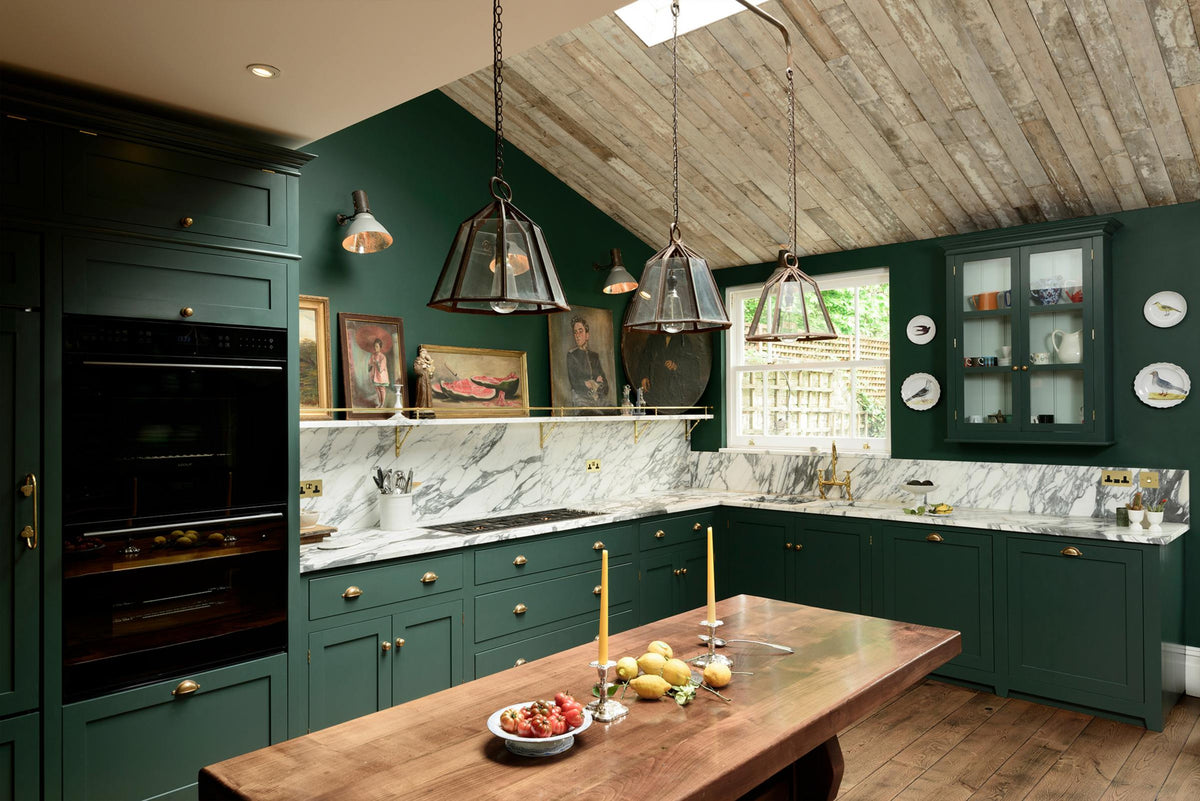 A lush green, classic English kitchen featuring an L-shaped sink and cooker, all complemented by Arabescato marble for the worktop, splashback, and countertop shelf. The kitchen includes full-height cabinets and a wall cupboard, creating a luxurious yet bohemian atmosphere. An angled wooden skylight window allows natural light to flow into the room, highlighting delicate bird-painted plates, a rich oak dining table, framed antique paintings, and industrial-style gas light fixtures on the walls.