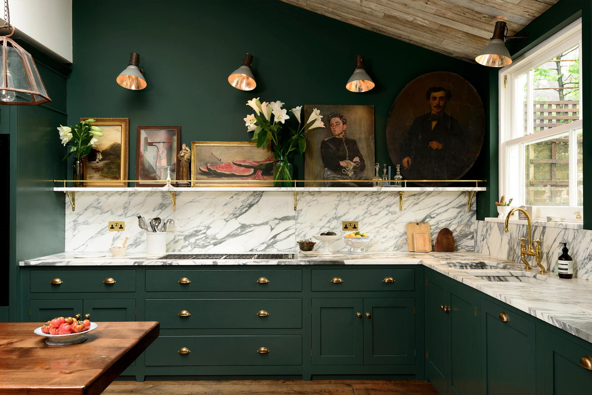 A lush green, classic English kitchen featuring an L-shaped sink and cooker, all complemented by Arabescato marble for the worktop, splashback, and countertop shelf. An angled wooden skylight window allows natural light to flow into the room, highlighting white lilies in glass vases, a rich oak dining table, framed antique paintings, and industrial-style gas light fixtures on the walls.