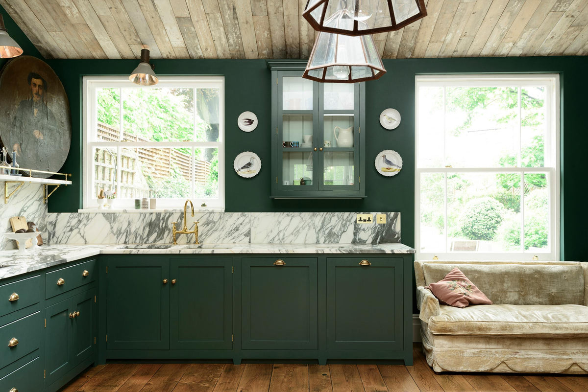 A classic English kitchen with lush greenery features an L-shaped sink and cooker, all enhanced by Arabescato marble for the worktop, splashback, countertop shelf, and wall cupboard, creating a luxurious yet bohemian atmosphere. An angled wooden skylight window allows natural light to flow into the room, highlighting delicate bird-painted plates, a cream velvet sofa, framed antique paintings, and industrial-style gas light fixtures on the walls.