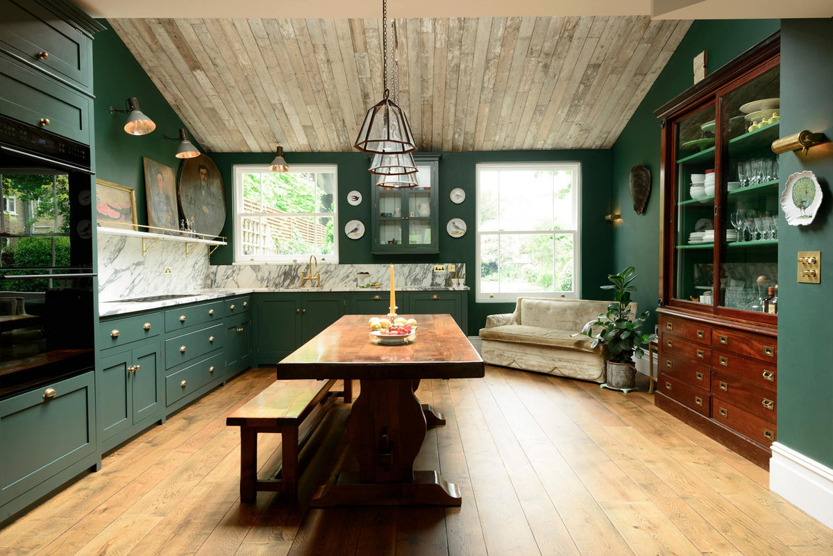 A lush green classic English kitchen featuring an L-shaped sink and cooker, all complemented by Arabescato marble for the worktop, splashback, and countertop shelf. The kitchen includes an antique glazed cabinet, a cream velvet sofa, full-height cabinets and a wall cupboard, creating a luxurious yet bohemian atmosphere. An angled wooden skylight window allows natural light to flow into the room, highlighting delicate bird-painted plates, a rich oak dining table, framed antique paintings, and industrial-styl