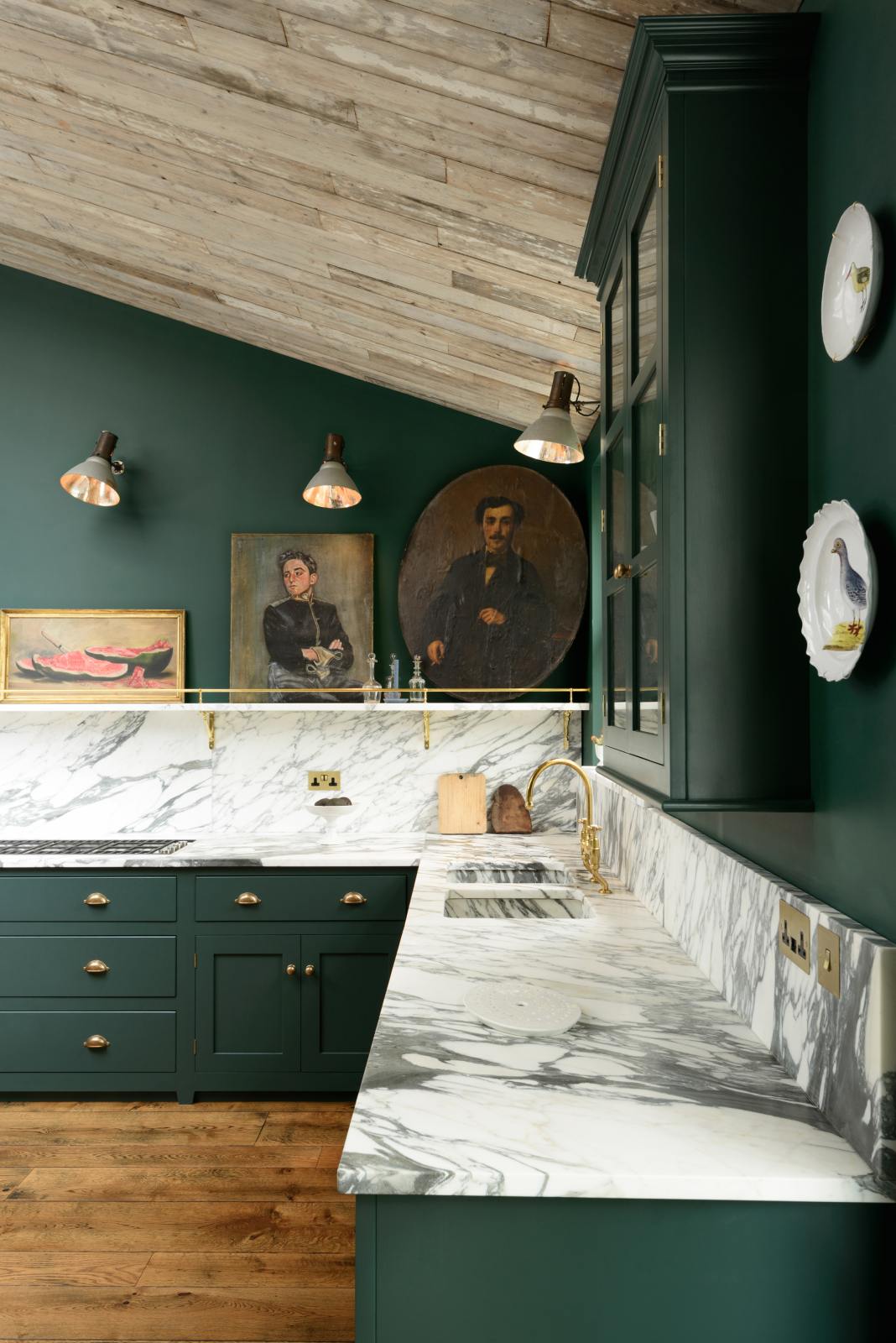 A quiet corner of a lush green, classic English kitchen featuring an L-shaped sink and cooker, all complemented by Arabescato marble for the worktop, splashback, countertop shelf, and a wall cupboard. An angled wooden skylight window allows natural light to flow into the room, highlighting delicate bird-painted plates, framed antique paintings, and industrial-style gas light fixtures on the walls.
