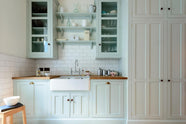 This pale-blue classic English galley kitchen features a sink run with a solid oak worktop, a double Belfast sink, a chrome Ionian tap, glazed wall cupboards, pantry cupboards, and shelves. Adorned with artisan ceramics, vintage containers, and simple platters, it creates a humble and practical kitchen that embraces vintage 80s aesthetics.