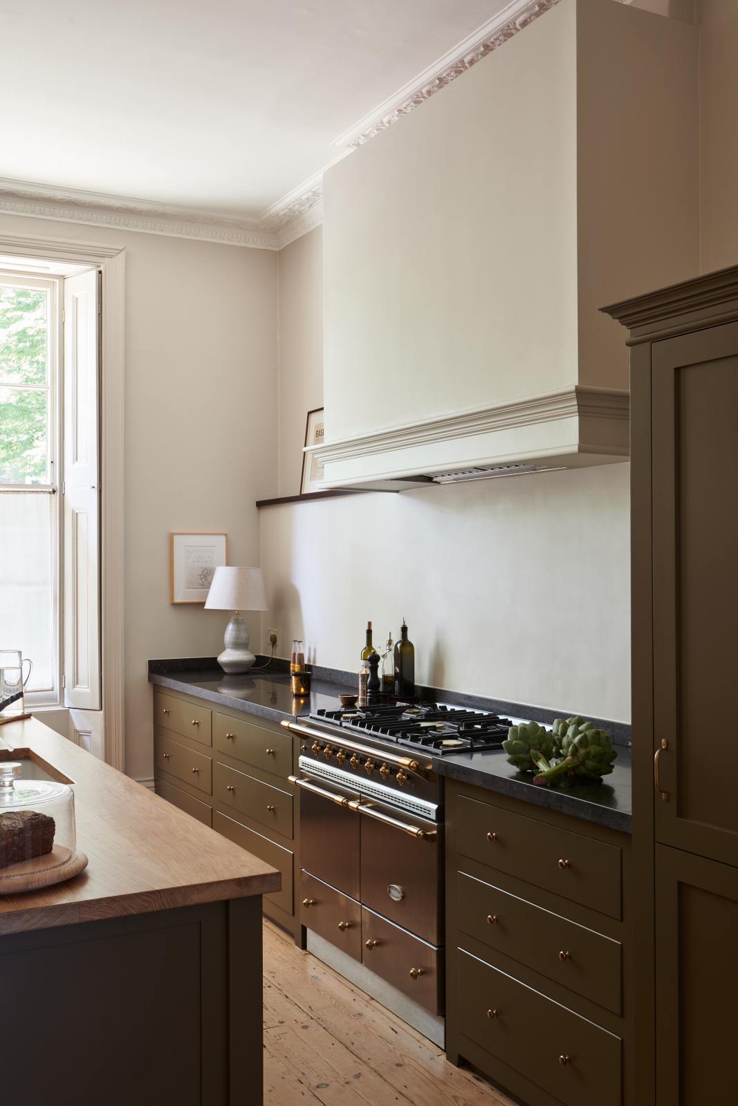 An oblique view of an elegant olive-brown classic kitchen features a cooker run topped with Belgian blue limestone, a kitchen island with a sink and a solid oak worktop, a shaker canopy, and a built-in shelf. The tall-ceilinged room is decorated with slender bottles of wine and vinegar, a pepper grinder, a vibrant green artichoke, and small, delicate framed illustrations.