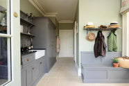 A functional muted-grey boot room features an L-shaped bench, shelves, a peg rail, a small sink run with a single Belfast Ionian sink, an antique silver Ionian tap, a black granite worktop, and full-height storage cupboards. Beautifully decorated with thick coats, a stack of hats, a woven shoulder bag, and a snowdrop potted plant, creating a practical and family-oriented place.