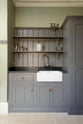 A closer look at a muted-grey boot room showcases a small sink run featuring a single Belfast Ionian sink, an antique silver Ionian tap, and a black granite worktop, complemented by shelves and full-height storage cupboards. Beautifully decorated with a vintage candelabra, a horseshoe, a beloved ceramic piece, and empty glass jars, it creates a practical and family-oriented space.