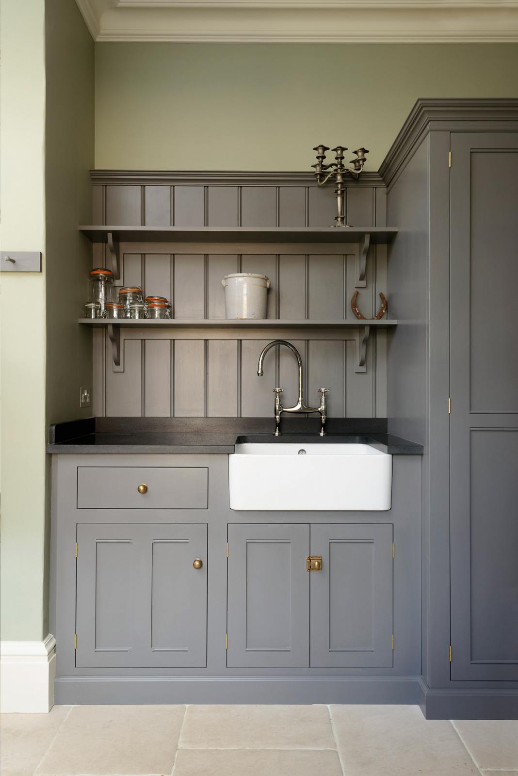 A closer look at a muted-grey boot room showcases a small sink run featuring a single Belfast Ionian sink, an antique silver Ionian tap, and a black granite worktop, complemented by shelves and full-height storage cupboards. Beautifully decorated with a vintage candelabra, a horseshoe, a beloved ceramic piece, and empty glass jars, it creates a practical and family-oriented space.
