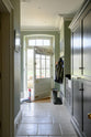 A functional muted-grey boot room features an L-shaped bench, shelves, a peg rail, a small sink run with a single Belfast Ionian sink, a black granite worktop, and full-height storage cupboards. Beautifully decorated with thick coats and muddy boots, this space leads into the back garden, creating a practical and family-oriented area.