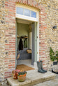 Passing a traditional British cottage with its textured wall, archway, and front door, you will find a functional, muted-grey boot room. This space features an L-shaped bench, a shelf with a peg rail, and a full-height storage cupboard. Beautifully decorated with thick coats, a stack of hats, a woven shoulder bag, muddy boots, and potted plants, it creates a practical and family-oriented space.