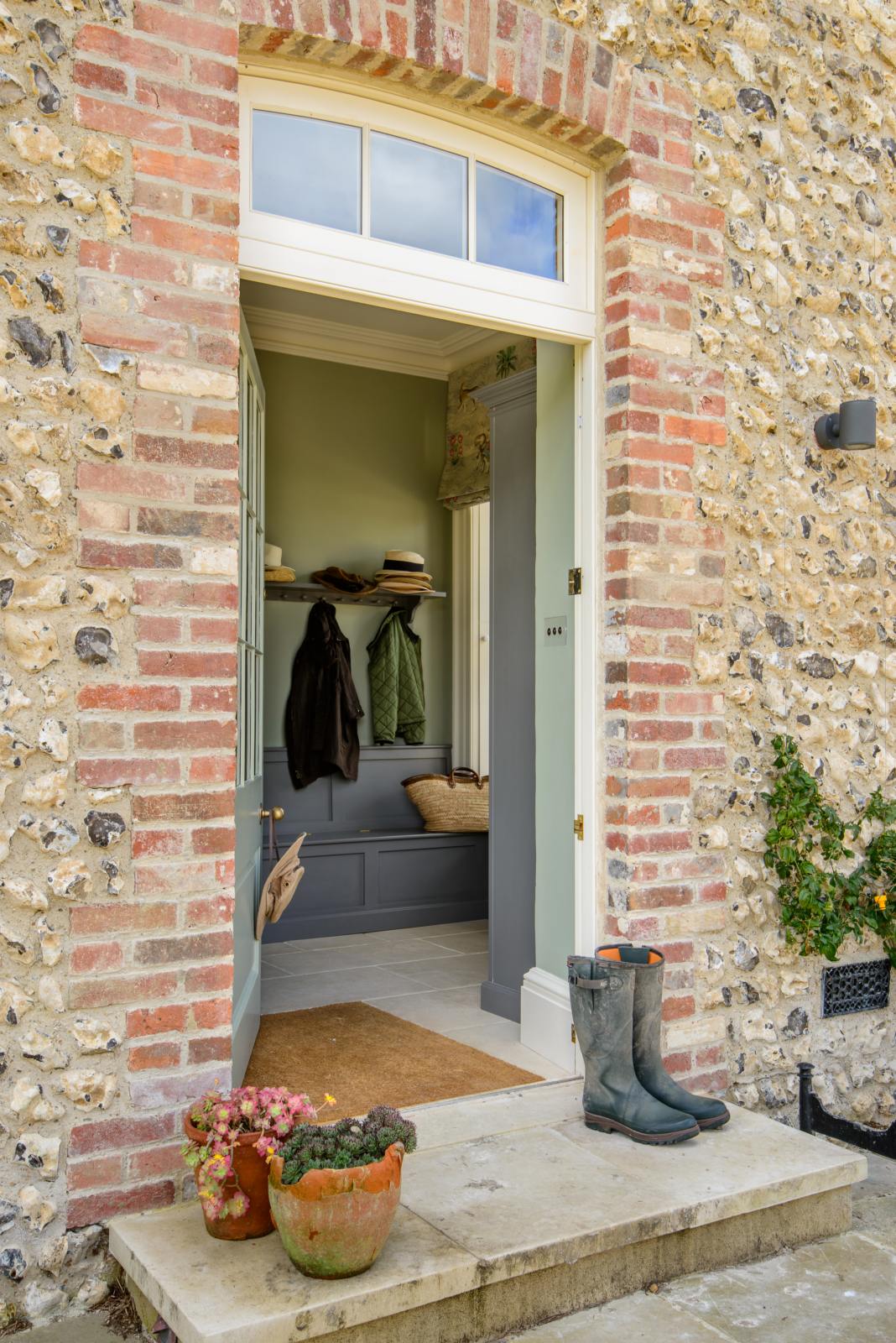 Passing a traditional British cottage with its textured wall, archway, and front door, you will find a functional, muted-grey boot room. This space features an L-shaped bench, a shelf with a peg rail, and a full-height storage cupboard. Beautifully decorated with thick coats, a stack of hats, a woven shoulder bag, muddy boots, and potted plants, it creates a practical and family-oriented space.