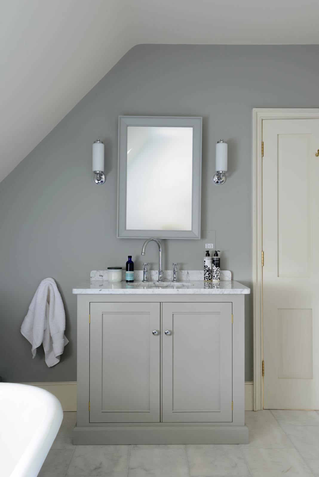 This simple and stylish muted-grey bathroom features a single sink stand with an Arabescato marble counter. Beautifully decorated with a mirror, silver wall lights, washing liquids, and a white towel, it creates a modern washstand and space.