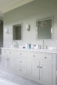 This simple and stylish muted-grey bathroom features a large washstand with two single sinks and an Arabescato marble counter. Beautifully decorated with mirrors, silver wall lights, washing liquids, and a crystal chandelier, it creates a modern and elegant space.