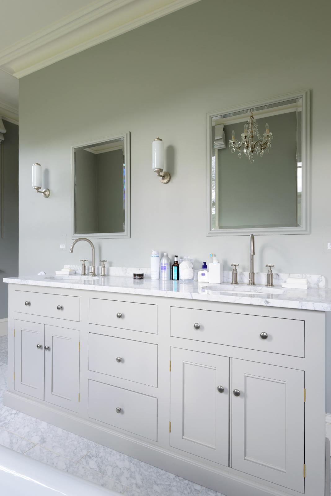 This simple and stylish muted-grey bathroom features a large washstand with two single sinks and an Arabescato marble counter. Beautifully decorated with mirrors, silver wall lights, washing liquids, and a crystal chandelier, it creates a modern and elegant space.