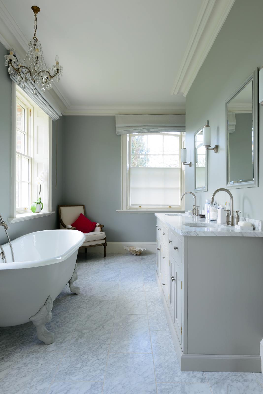 This simple and stylish muted-grey bathroom features a large washstand with two single sinks, an Arabescato marble counter, and a white bathtub. Beautifully decorated with mirrors, silver wall lights, washing liquids, and a crystal chandelier, it creates a modern and elegant space.