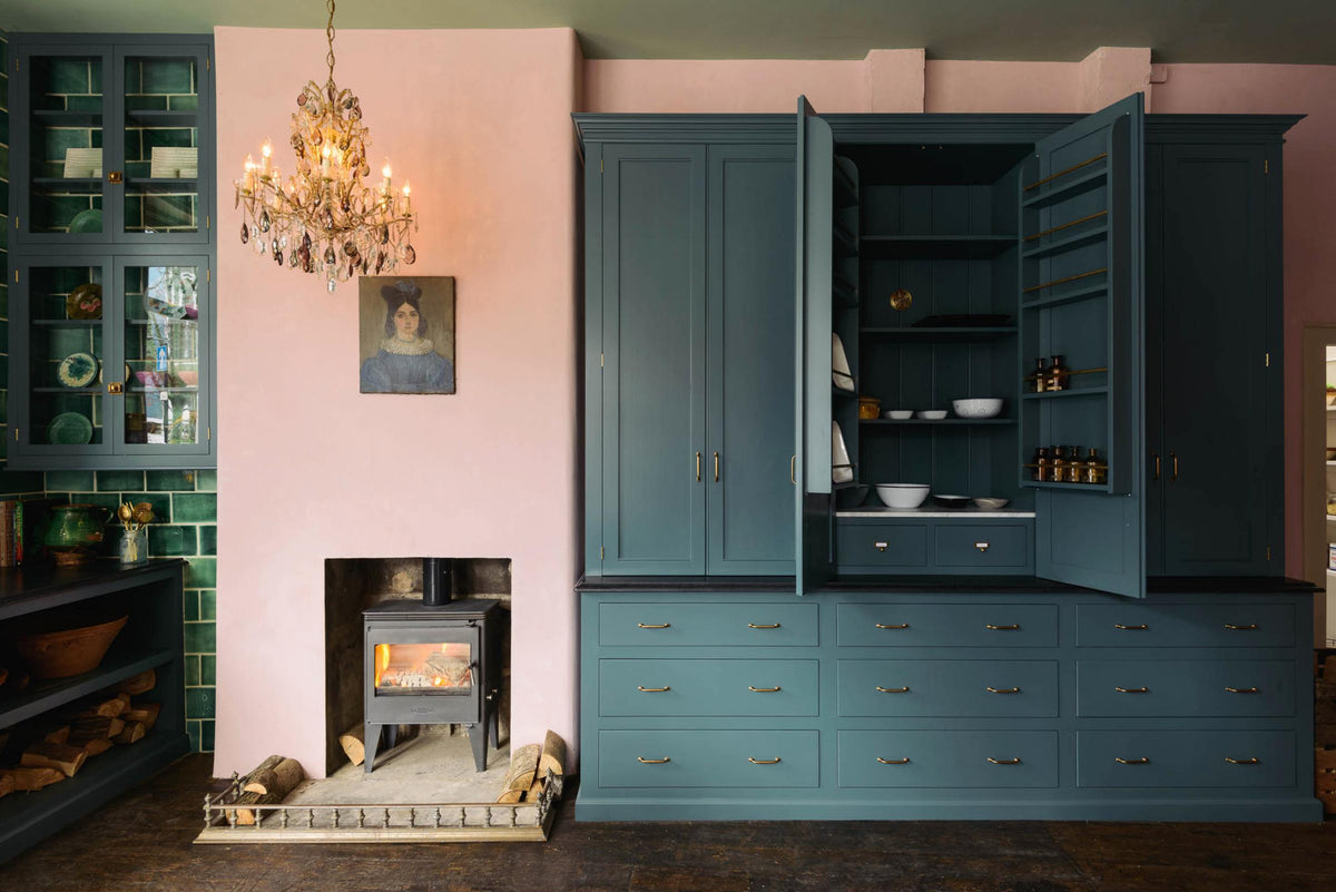 The playful pastel yet classic kitchen features a large Clerkenwell blue full-height pantry cupboard with a black granite worktop, emerald green splashback tiles, and a pink chimney with a small iron wood stove. Decorated with chopped firewood, vintage ceramics, a small oil painting portrait and a crystal chandelier, this kitchen has both functionality and personality.