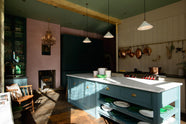 A dramatic image inside the deVOL St John's Square, London showroom. The spacious room shows a playful pastel yet classic kitchen features a mushroom-coloured cooker run, aged brass hanging rails, and a pot filler. A long kitchen island with a breakfast nook and deVOL bum stools adds charm. Finally, a Clerkenwell blue sink run with a black granite worktop, and emerald green splashback tiles. Decorated with copper pots, vintage ceramics, and fresh produce, this kitchen has both functionality and personality.