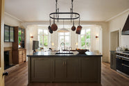 A spacious and sophisticated olive green classic kitchen with a large kitchen island, Belgian blue limestone worktop with a single sink and antique silver Ionian tap. Above, a deVOL pot hanger takes centre stage, while three large windows flood the kitchen and dining room with lovely light.
