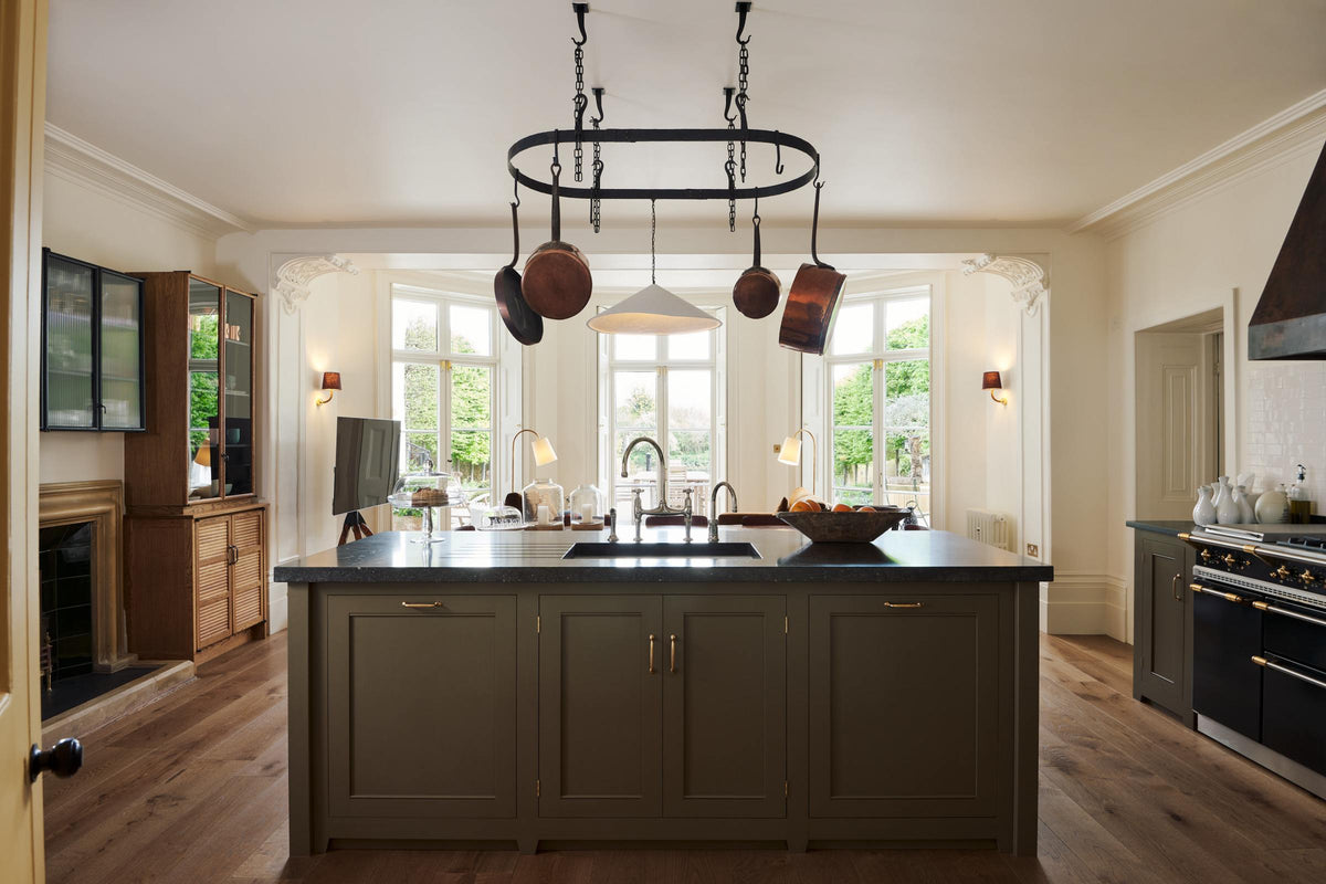A spacious and sophisticated olive green classic kitchen with a large kitchen island, Belgian blue limestone worktop with a single sink and antique silver Ionian tap. Above, a deVOL pot hanger takes centre stage, while three large windows flood the kitchen and dining room with lovely light.