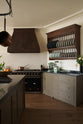 A spacious olive-green classic kitchen features a large island with a Belgian blue limestone worktop, a single sink, and an antique silver Ionian tap. Above, a deVOL pot hanger takes centre stage. The classic cooker run showcases a large black gas stove, a substantial canopy, and an heirloom plate rack displaying colour-coordinated muted greens and dark blue ceramic plates.