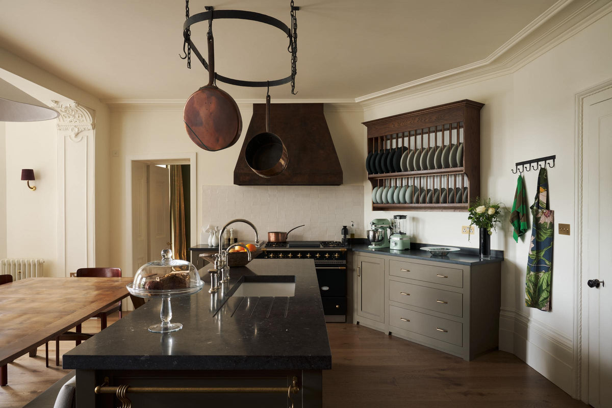 A spacious olive-green classic kitchen features a large island with a Belgian blue limestone worktop, a single sink, and an antique silver Ionian tap. Above, a deVOL pot hanger takes centre stage. The classic cooker run showcases a large black gas stove, a substantial canopy, white splashback tiles, and an heirloom plate rack displaying colour-coordinated muted greens and dark blue ceramic plates.