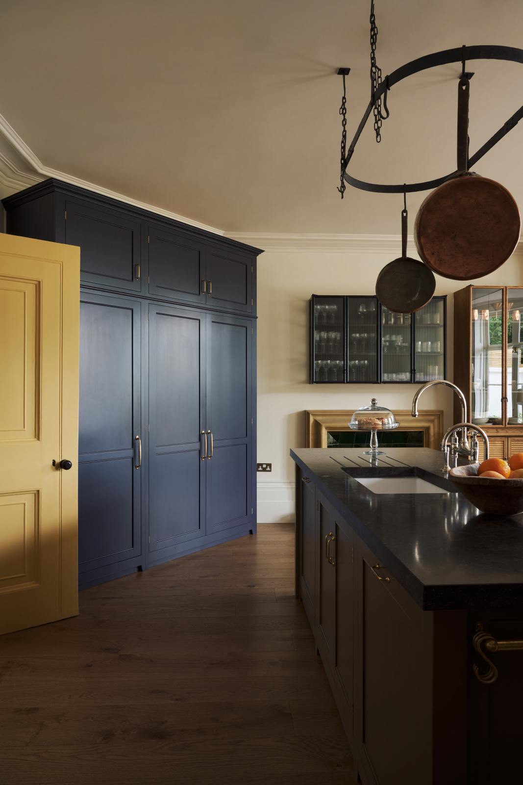 The spacious olive-green kitchen has a large island with a Belgian blue limestone worktop, a single sink, and an antique silver Ionian tap, topped by a deVOL pot hanger. It features a full-height pantry cabinet, Haberdasher wall cupboards, and a freestanding pantry, all surrounding a grand fireplace framed in rusty gold and emerald green tiles.