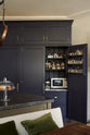 A detailed view of a large, dark blue, full-height pantry cabinet with an open door reveals its features: a spice rack, a combination of open and closed cabinets, and a marble countertop displaying a variety of dried ingredients, along with a microwave and toaster. In front of the pantry stands an olive-green kitchen island topped with Belgian blue limestone.