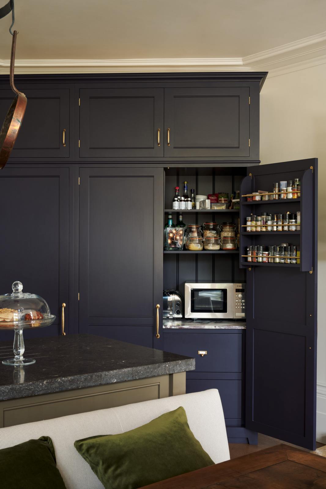 A detailed view of a large, dark blue, full-height pantry cabinet with an open door reveals its features: a spice rack, a combination of open and closed cabinets, and a marble countertop displaying a variety of dried ingredients, along with a microwave and toaster. In front of the pantry stands an olive-green kitchen island topped with Belgian blue limestone.