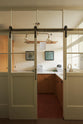 A cream sliding door leads to a scullery yellow laundry room featuring an L-shaped sink with a white marble countertop and a stunning aged brass Ionian faucet. Neatly stacked white towels are arranged in layers, and two framed artworks adorn the walls. In the centre of the ceiling hangs a deVOL Gaselier light, while a deVOL Laundry Maid, with a towel draped over it, is positioned behind.